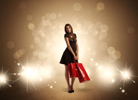 A beautiful elegant woman in black standing with red shopping bags in front of brown background and bright glowing lights concept