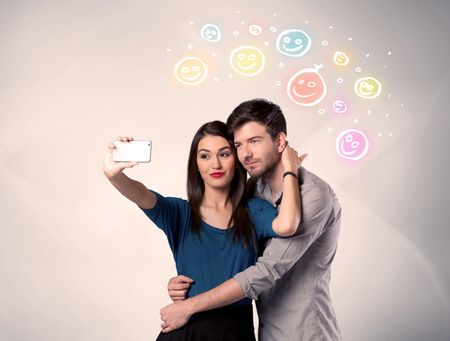 A cheerful young couple taking selfie photo with mobile phone and colorful happy smiley faces illustration above them concept