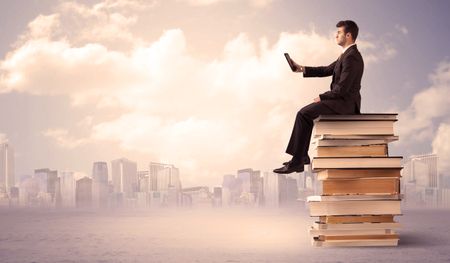 A serious student with laptop tablet in elegant suit sitting on a stack of books in front of cityscape with clouds