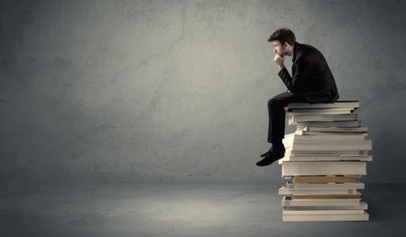 A serious student in elegant suit sitting on a stack of books in front of dark grey background