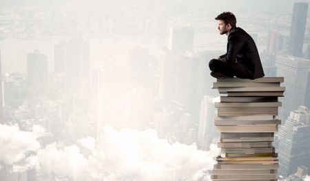 A serious businessman in elegant suit sitting on a pile of giant books in front of a grey city scape with clouds, fog