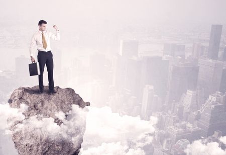 A young successful happy businessman standing confident on stone cliff in the clouds looking at the city scape from above concept