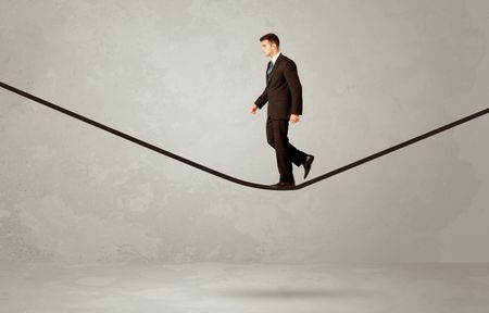 An elegan businessman in suit balancing on a tight rope with a briefcase in front of grey urban wall background environment concept