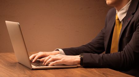 An office worker in elegant suit sitting at desk, typing on portable laptop with empty brown wall background