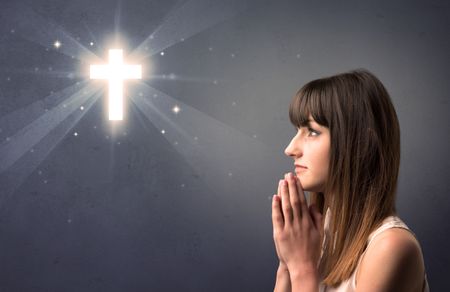 Young woman praying on a grey background with a shiny cross above her