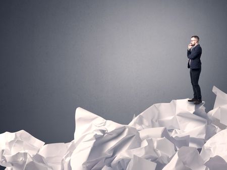 Thoughtful young businessman standing on a pile of crumpled paper with a grey background