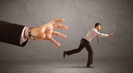 Young miniature businessman running from a big hand with grey background