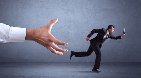 Young miniature businessman running from a big hand with blueish grey background