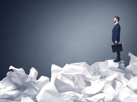 Thoughtful young businessman standing on a pile of crumpled paper with a blueish grey background