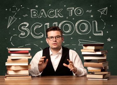 A young teacher in glasses sitting at classroom desk with pile of books in front of blackboard saying back to school drawing concept.