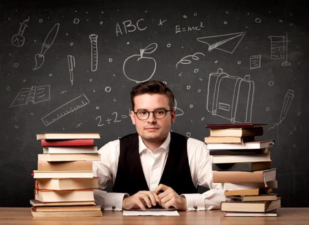 A passionate young teacher sitting at school desk with pile of books in front of blackboard drawn full of back to school items concept.