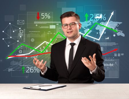 Young handsome businessman sitting at a desk with stocks and progress charts behind him