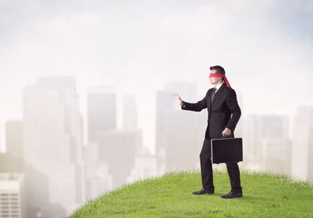 Young blindfolded businessman steps on a a patch of grass with a city in the background 