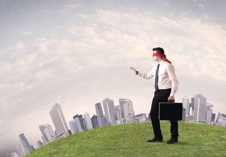 Young blindfolded businessman steps on a a patch of grass with a grey buildings in the background