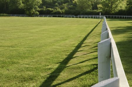 Edge of racetrack infield, with continuous white fence with symmetrical shadow, on late summer afternoon at equestrian training center