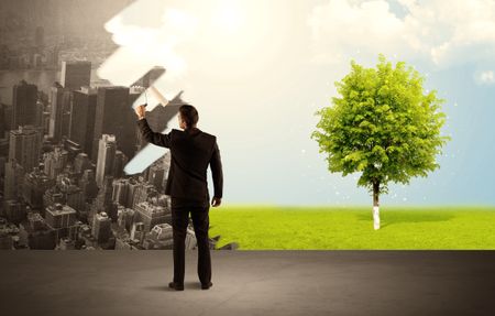 Businessman in elegant suit standing with his back, holding a roller and transforming the grey city landscape into green tree in bright nature environment concept