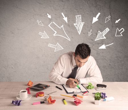 A young businessman sitting at an office desk and working on paperwork with drawn arrows pointing at his head concept