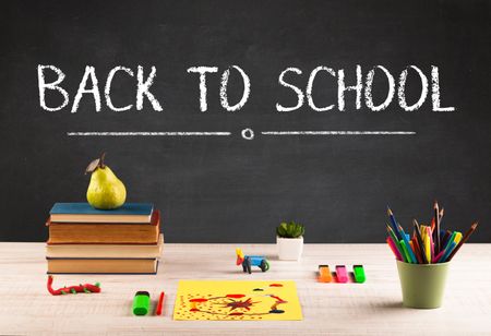 Back to school concept with writing on blackboard in capital letters and a desk with papers, fruit