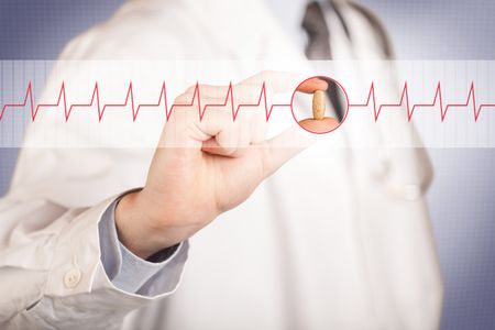 A male doctor in white coat with a stethoscope on one shoulder holding a pill between his fingers focused on a heartbeat graph