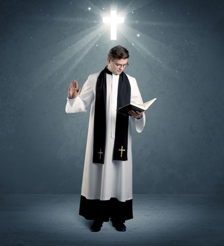 A young caucasian priest with deep faith blessing with the holy bible in his hand in front of blue wall background with illustrated glowing cross concept.