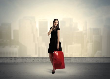 Attractive lady in black holding red shopping bags standing in front o urban landscape with tall buildings concept