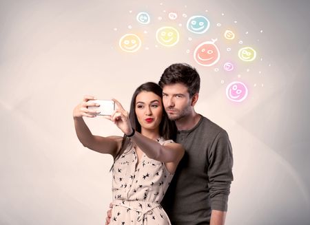 A cheerful young couple taking selfie photo with mobile phone and colorful happy smiley faces illustration above them concept
