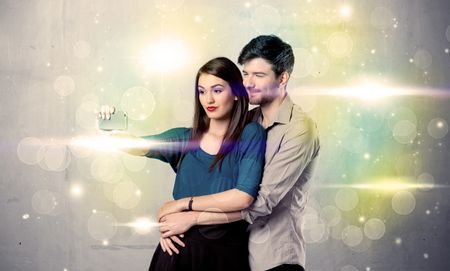 A fresh happy couple taking selfie photo with mobile phone in front of colorful lights glitter wall background concept