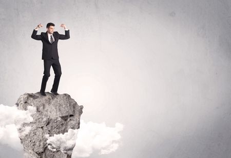 A happy successful businessman standing on a stone cliff with clouds in front of clear empty grey background concept