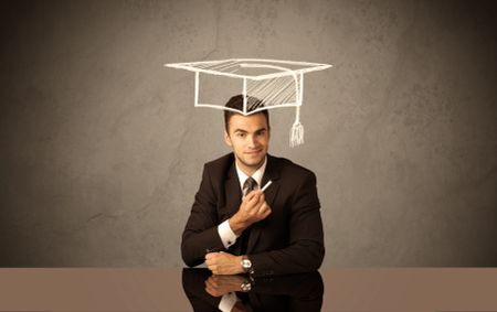 An elegant, successful university student drawing himself a square academic mortarboard cap with a chalk in front of grey wall background concept