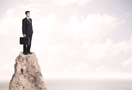 Successful sales person with brief case standing on top of a mountain cliff edge looking above the landscape between the clouds