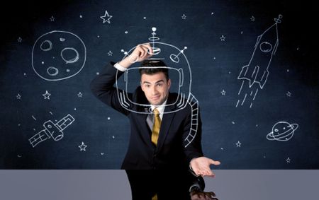 A young happy businessman in elegant suit drawing helmet, satellite and rocket in empty space with a chalk illustration concept