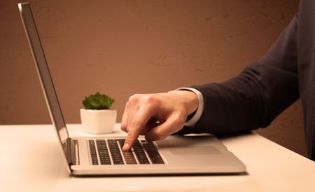 An office worker in elegant suit sitting at desk, typing on portable laptop with empty brown wall background