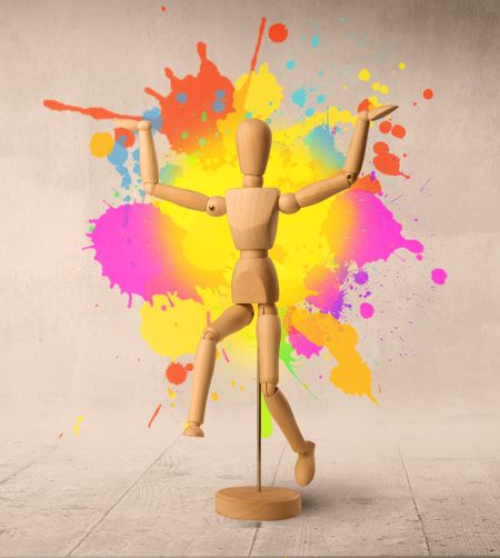 Wooden mannequin posed in front of a greyish background with colorful splashes behind it