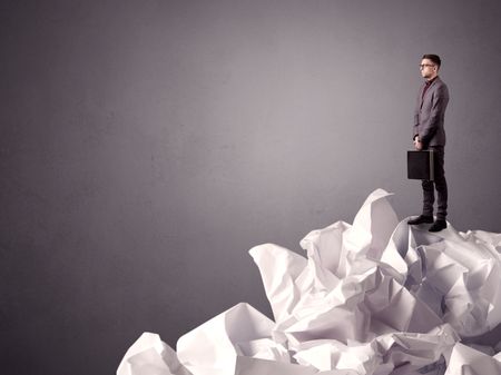Thoughtful young businessman standing on a pile of crumpled paper with a grungy grey background