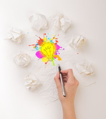 Female hand next to a few crumpled paper balls drawing a colorful lightbulb