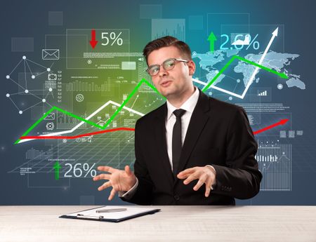Young handsome businessman sitting at a desk with stocks and progress charts behind him