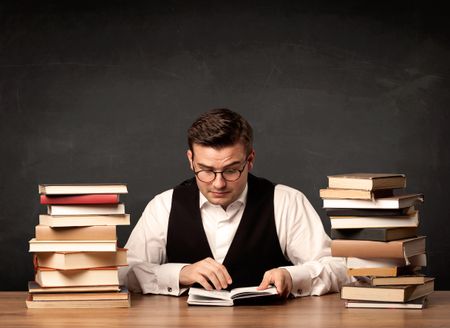 A young teacher in glasses sitting at classroom desk with pile of books in front of clean blackboard back to school concept.