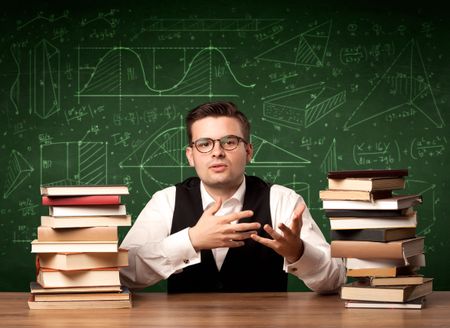 A young passionate male teacher sitting at school desk, reading a book, with area algorythm calculations and numbers on the blackboard concept.