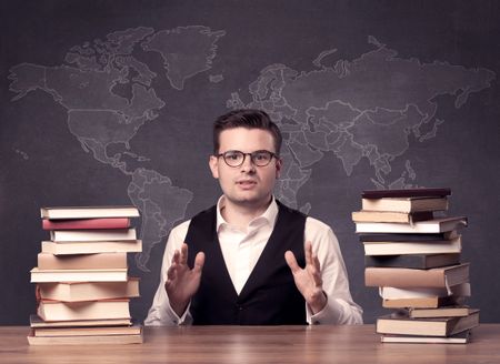 A young ambitious geography teacher in glasses sitting at classroom desk with pile of books in front of world map drawing on blackboard, back to school concept.