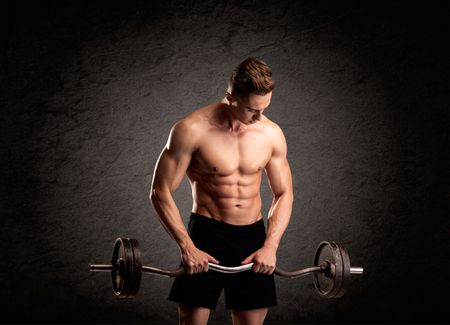 A handsome fitness guy lifting weight and showing his strong muscles in front of a black urban concrete wall concept