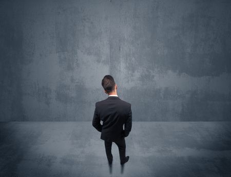 A young businessman with briefcase standing in blank empty space facing a grey urban wall, scratching his head concept.