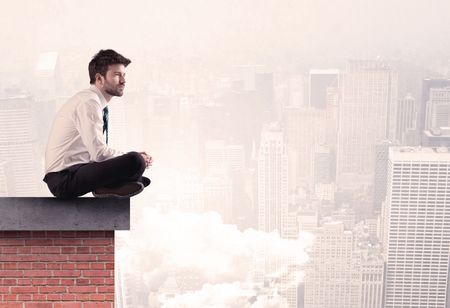 An elegant businessman in modern suit sitting on the top of a brick building, looking over the cityscape with clouds concept