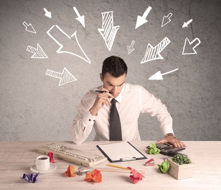 A young businessman sitting at an office desk and working on paperwork with drawn arrows pointing at his head concept