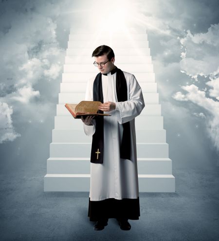 A young religious happy priest standing in front of the stairway to heaven concept with clouds and bright lights coming from above.