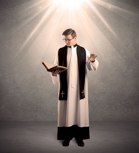 A male religious young priest in black and white dress giving his blessing, holding the holy bible while being illuminated from strong light beams coming from above concept