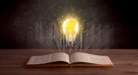 Shiny alphabet letters and yellow lightbulb hovering over open book 