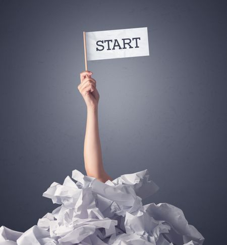 Female hand emerging from crumpled paper pile holding a white flag with start written on it