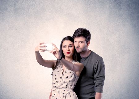 A young couple in love taking selfie with a mobile phone in the handsome guy's hand in front of an empty clear grey wall background concept