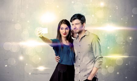 A fresh happy couple taking selfie photo with mobile phone in front of colorful lights glitter wall background concept