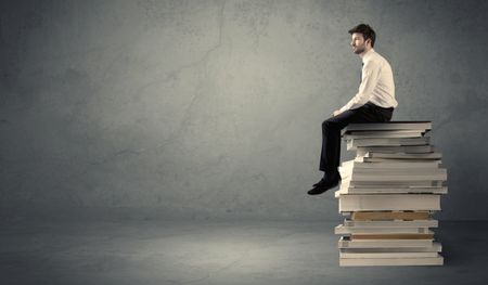 A serious student in elegant suit sitting on a stack of books in front of dark grey background